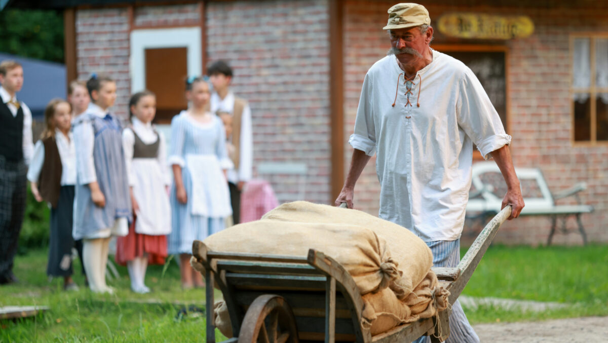 Ein älterer Mann in historischer Kleidung schiebt eine hölzerne Schubkarre, die mit Leinensäcken gefüllt ist, als Teil der Darß-Festspiele, während sich eine Gruppe von Menschen in historischen Kostümen in der Nähe eines Backsteingebäudes im Hintergrund versammelt.