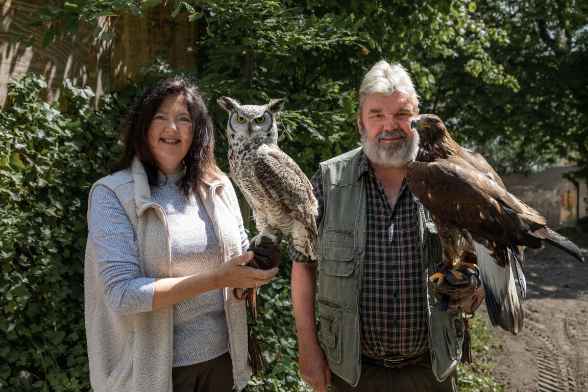 Eine lächelnde Frau (Lisa Walter) und ein lächelnder Mann (Volker Walter) stehen bei den Störtebeker Festspielen im Freien und halten jeweils einen Raubvogel in der Hand; die Frau hält eine Eule, der Mann einen großen braunen Adler. Grünes Laub und Sonnenlicht bilden eine natürliche Kulisse.