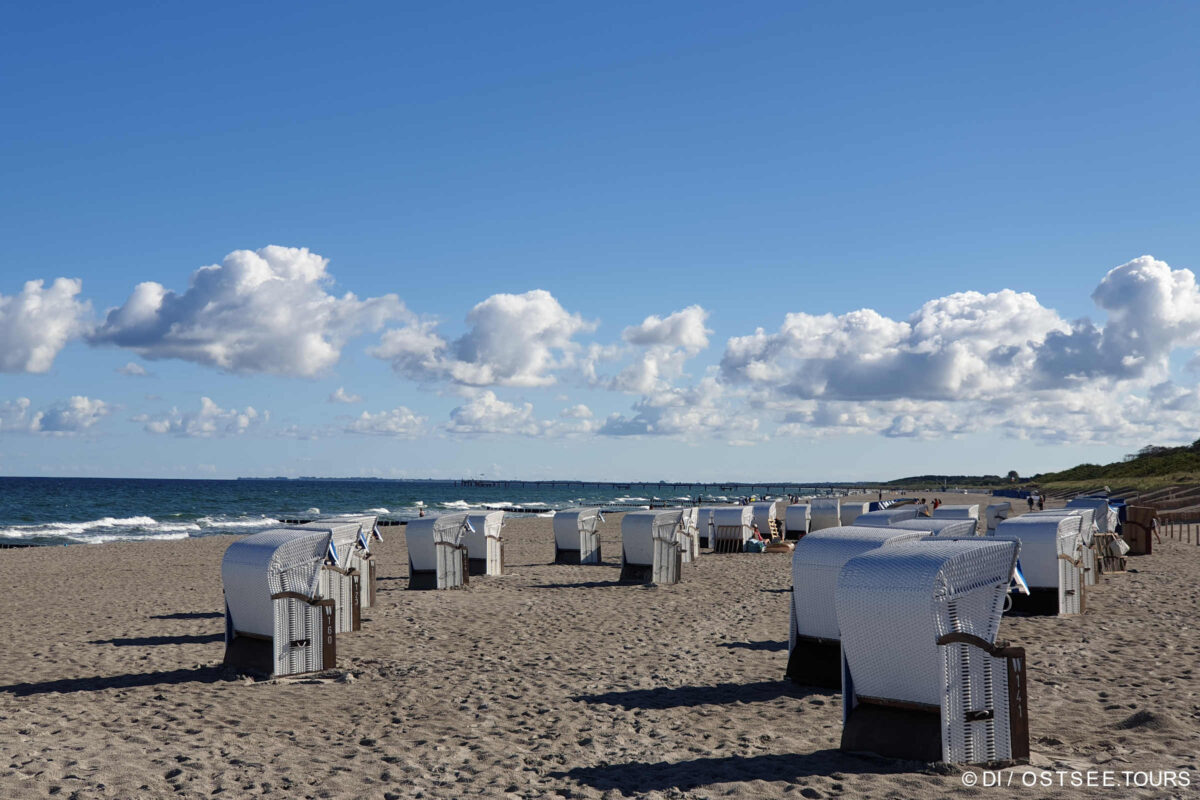 Strandkörbe stehen am Sandstrand des Ostseeheilbades Graal-Müritz unter einem blauen Himmel mit vereinzelten Wolken. Links erstreckt sich das Meer, rechts sind grasbewachsene Dünen zu sehen.
