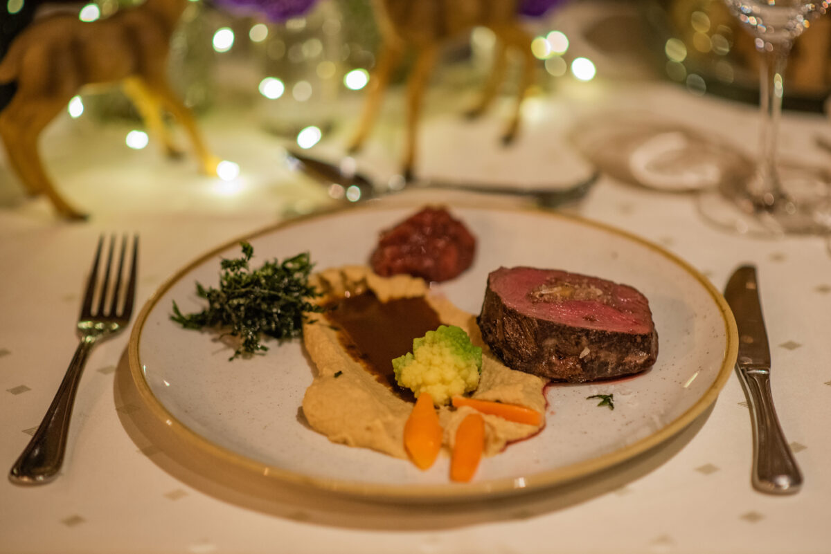 Ein von den Wildwochen inspiriertes Gourmet-Menü, bestehend aus einer Scheibe Rindfleisch, brauner Soße, cremigem Püree, grünem und orangefarbenem Gemüse sowie einem Klecks rotem Chutney, serviert auf einem festlich gedeckten Tisch vor dem Hintergrund festlicher Lichter – perfekt, um die kulinarischen Köstlichkeiten von Usedom zu genießen.